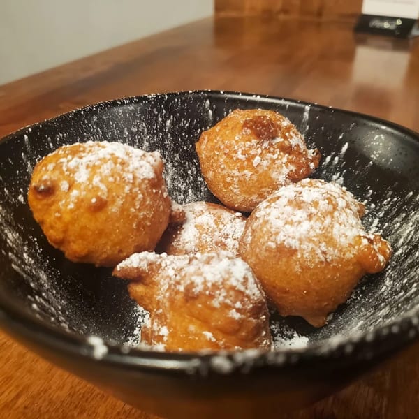 Five golden-brown fried dough balls, dusted with powdered sugar, in a black bowl on a wooden table.