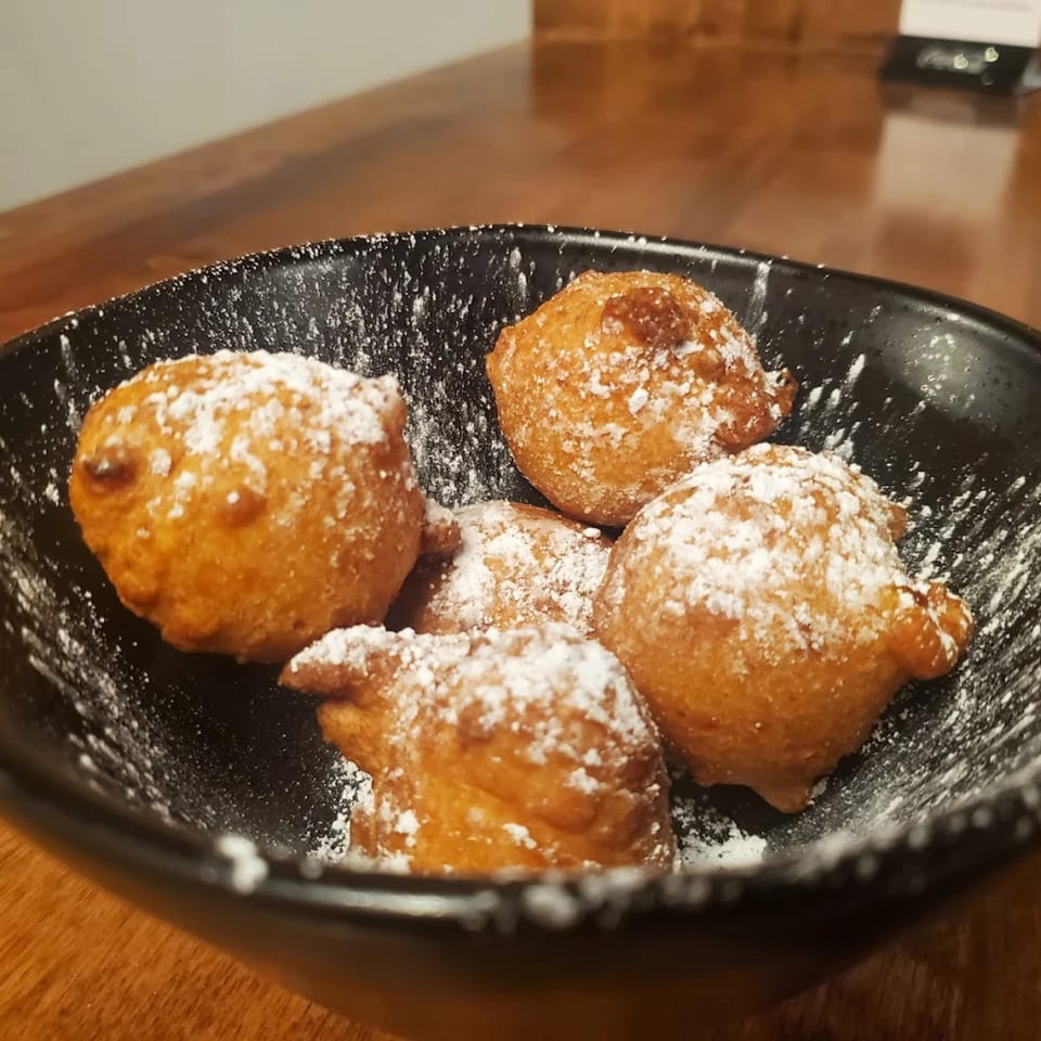 Five golden-brown fried dough balls, dusted with powdered sugar, in a black bowl on a wooden table.