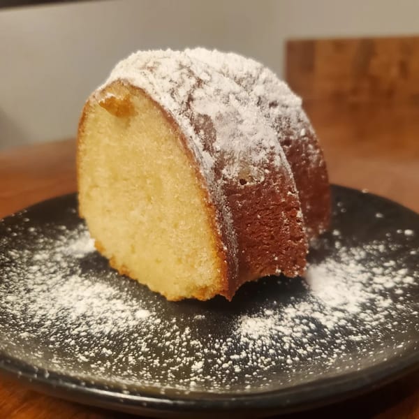 A slice of golden-brown pound cake, dusted with powdered sugar, rests on a dark plate.