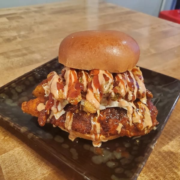 A Nashville Hot Chicken sandwich with crispy fried chicken, coleslaw, and sauces on a dark plate.