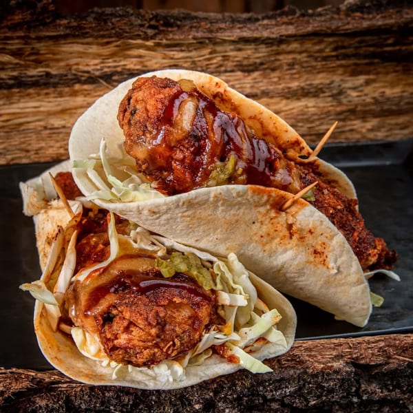 Two Fire Nashville Hot Chicken tacos with fried chicken, slaw, and sauce on a rustic wooden surface.
